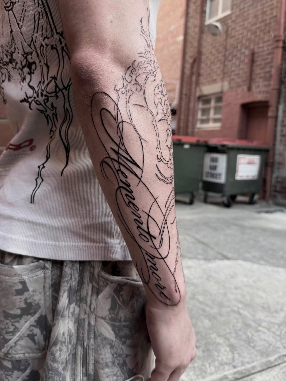 Forearm tattoo of the words “Memento Mori” in flourished script over ornamental linework, photographed in a Perth laneway